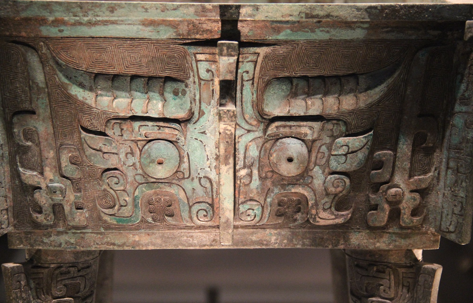 The frontal taotie animal-mask cast in relief on the body of a Western Zhou bronze ding cauldron — a symmetrical face with two great raised bosses for eyes, curling horns and quills above, and a split profile-dragon body flanking a central ridge, all over a fine leiwen (thunder-pattern) ground; two legs of the cauldron show below.